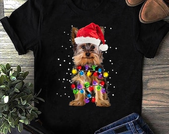 Może przedstawiać: Czarny t-shirt z Yorkshire Terrierem w czapce Świętego Mikołaja i lampkami choinkowymi. Pies jest brązowy i brązowy z czerwonym kapeluszem. Światła są wielokolorowe. Białe kropki sugerują śnieg.