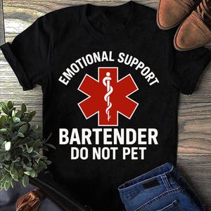 May include: Black t-shirt featuring the text "EMOTIONAL SUPPORT BARTENDER DO NOT PET" in white, with a red medical symbol. The shirt is made of a soft material and is suitable for casual wear.