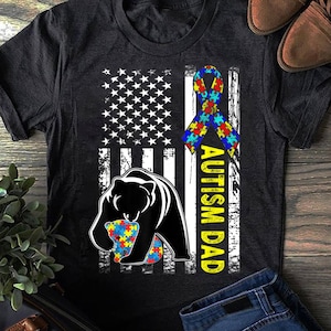 May include: Black t-shirt with a distressed American flag design. The flag has a blue and yellow ribbon with puzzle pieces, and a black bear holding a puzzle piece. The text "Autism Dad" is written vertically on the right side of the flag.