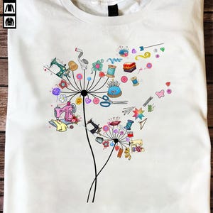 May include: White t-shirt featuring a colorful dandelion design composed of sewing tools and accessories. The design includes sewing machines, spools of thread, scissors, and other craft supplies. The graphic is a whimsical representation of the sewing and crafting theme.
