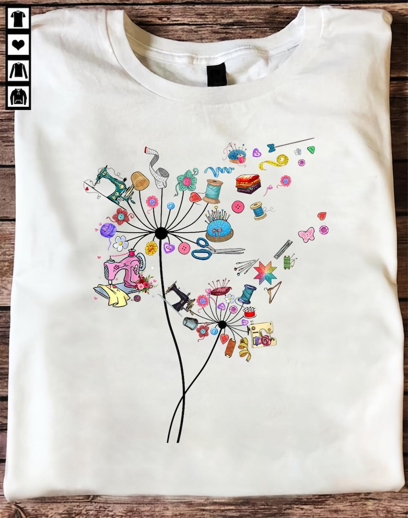 May include: White t-shirt featuring a colorful dandelion design composed of sewing tools and accessories. The design includes sewing machines, spools of thread, scissors, and other craft supplies. The graphic is a whimsical representation of the sewing and crafting theme.