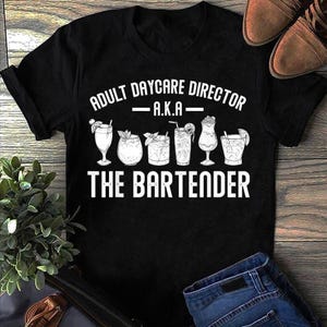 Puede incluir: Camiseta negra con texto blanco: "ADULT DAYCARE DIRECTOR - A.K.A - THE BARTENDER". El diseño incluye ilustraciones de cócteles. La imagen también muestra un par de zapatos marrones y unos vaqueros azules.