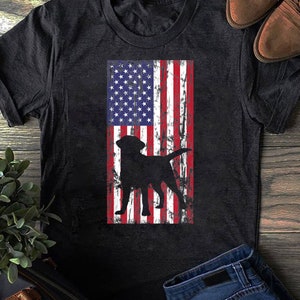May include: A black t-shirt featuring a distressed American flag design with a black silhouette of a Labrador Retriever dog.