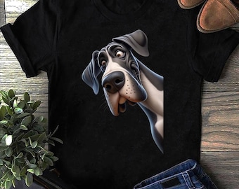 Può includere: T-shirt nero con un'illustrazione a cartoni animati di un Alano sorpreso. Il cane è principalmente bianco e nero, con un'espressione dettagliata. La maglietta è abbinata a jeans blu e scarpe marroni.