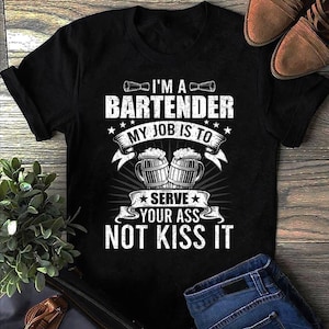 Puede incluir: Camiseta negra con un gráfico blanco que dice "I'M A BARTENDER MY JOB IS TO SERVE YOUR ASS NOT KISS IT". El diseño incluye imágenes de jarras de cerveza y estrellas. La camiseta se muestra sobre una superficie de madera.