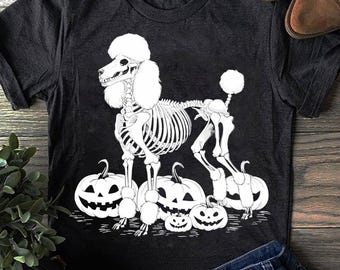 May include: Dark gray t-shirt featuring a white skeleton poodle design. The poodle is surrounded by white jack-o'-lanterns. A fun Halloween themed graphic tee.