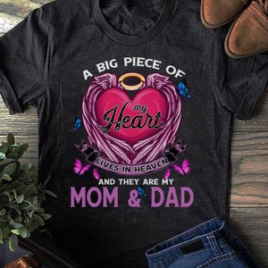 May include: Dark gray t-shirt with a heart-shaped graphic. The heart is pink with purple wings and the words "my Heart" in cursive. The text reads "A BIG PIECE OF my Heart LIVES IN HEAVEN AND THEY ARE MY MOM & DAD."