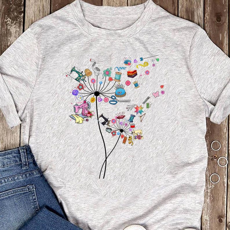 May include: A light gray t-shirt featuring a colorful dandelion design. The dandelion's seeds are replaced with sewing-themed elements like sewing machines, spools of thread, and scissors. The design is in various colors, including pink, green, and blue.