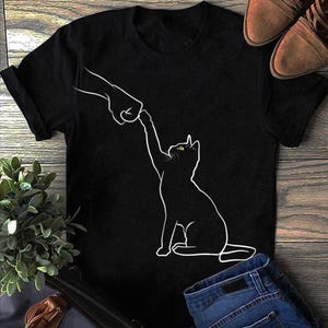 Puede incluir: Camiseta negra con un contorno blanco de un gato que alcanza una mano. El gato tiene ojos amarillos. La camiseta está sobre una superficie de madera con zapatos marrones y vaqueros azules.