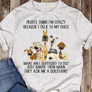 May include: A light gray t-shirt featuring a graphic of several cartoon dogs. The text on the shirt reads "PEOPLE THINK I'M CRAZY BECAUSE I TALK TO MY DOGS" and "WHAT AM I SUPPOSED TO DO? JUST IGNORE THEM WHEN THEY ASK ME A QUESTION?"