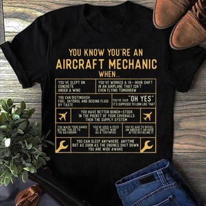 以下が含まれることがあります： 「You know you're an Aircraft Mechanic when...」という金色のグラフィックがプリントされた黒いTシャツ。航空機の整備士であることのユーモラスな兆候がいくつかリストされています。
