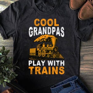 May include: Dark grey t-shirt with the text "COOL GRANDPAS PLAY WITH TRAINS" in white and orange. A detailed illustration of a vintage train is featured below the text. The shirt is made of a soft material.