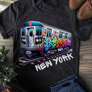 Puede incluir: Una camiseta gris carbón con un gráfico de un tren de metro cubierto de arte de grafiti colorido. El tren está posicionado sobre vías de ferrocarril y el texto NEW YORK está impreso en letras mayúsculas blancas bajo la ilustración.