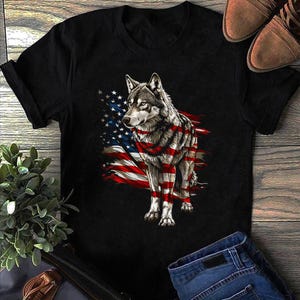 May include: Black t-shirt featuring a detailed illustration of a wolf with an American flag design. The wolf is depicted with the flag's stripes and stars integrated into its fur, creating a patriotic and artistic design.
