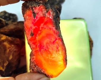 May include: A rough, irregularly shaped stone with a vibrant, glowing red and orange interior. The stone has a dark, textured exterior and is held by a hand. The overall appearance suggests a natural, possibly mineral specimen.