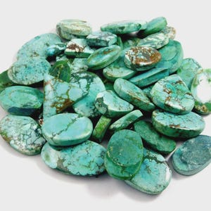 May include: A collection of turquoise gemstones in different shapes. The stones show a range of blue-green shades with brown matrix patterns. The gemstones are oval, round, and irregular, with a polished finish.
