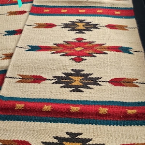 May include: A white woven rug with a geometric pattern in shades of red, brown, black, and teal. The pattern features arrows and diamond shapes.