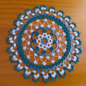 Puede incluir: Un encaje de ganchillo blanco y azul con un borde verde. El encaje tiene un diseño floral en el centro.