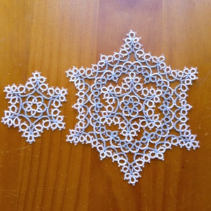 Könnte beinhalten: Zwei weiße und blaue gehäkelte Schneeflocken auf einer Holzoberfläche. Die größere Schneeflocke ist im Hintergrund und die kleinere Schneeflocke im Vordergrund.