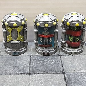 May include: Three miniature sci-fi themed containers. Each cylindrical container has a silver top and base, with yellow accents. One container features a gold and yellow design, another contains a gray alien, and the last has green tentacles.