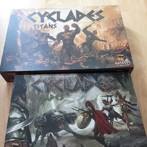 Puede incluir: Dos cajas de juego para el juego de mesa Cyclades. La caja superior se titula "Cyclades: Titans" y presenta a un guerrero con una espada y un escudo. La caja inferior se titula "Cyclades: Hades" y presenta a un guerrero con un escudo y una espada luchando contra un perro de tres cabezas.