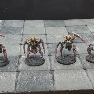 Może przedstawiać: Cztery miniaturowe figurki fantasy, z których każda przedstawia humanoidalne stworzenie o cechach owada. Figurki mają złote głowy, brązowe torsy i fioletowe kończyny. Ustawione są na szarej, kafelkowanej powierzchni, sugerującej grę planszową.