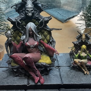 May include: Two miniature figures of women seated on thrones. The woman on the left is wearing a red dress and has long white hair. The woman on the right is wearing a green dress and has long blonde hair. Both figures are seated on thrones made of dark wood and have skulls on their heads.