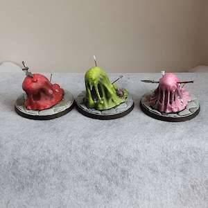 以下が含まれることがあります： 緑、ピンク、赤の粘液モンスターのミニチュアフィギュア3体。武器が刺さっています。フィギュアは丸い台座の上にあります。