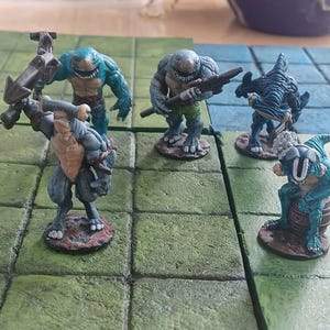 May include: Five painted miniature figurines of blue and green aquatic creatures, standing on a green and brown tiled surface. The figurines are holding weapons and are posed in a fighting stance.