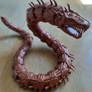 May include: A detailed, bronze-colored, articulated serpent figurine. The creature has a segmented body, a detailed head with visible teeth, and a spiked dorsal ridge. The design suggests a fantasy or mythological theme.