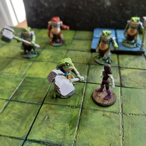 Puede incluir: Una figura en miniatura de rana verde sosteniendo un martillo plateado, de pie sobre una baldosa cuadrada verde. Hay otras figuras en miniatura en el fondo, incluyendo una rana roja, una rana verde y una figura humana morada.