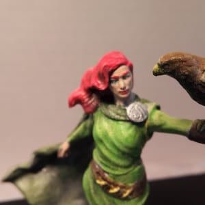 May include: A red-haired woman figurine with a green cloak and dress, holding a brown hawk figurine on her outstretched arm.