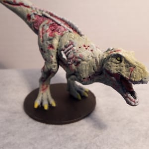 Pode incluir: Uma miniatura detalhada de um dinossauro zumbi, pintada em tons de cinza, branco e vermelho. O dinossauro tem uma cabeça grande com uma boca aberta e dentes afiados. A miniatura está colocada em uma base marrom.