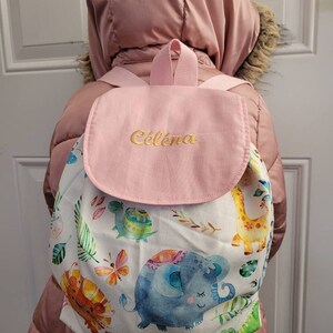 以下が含まれることがあります： ピンクと白のバックパックで、フラップには「Celena」という名前が刺繍されています。バックパックは白い背景にカラフルなジャングル動物のプリントが施されています。