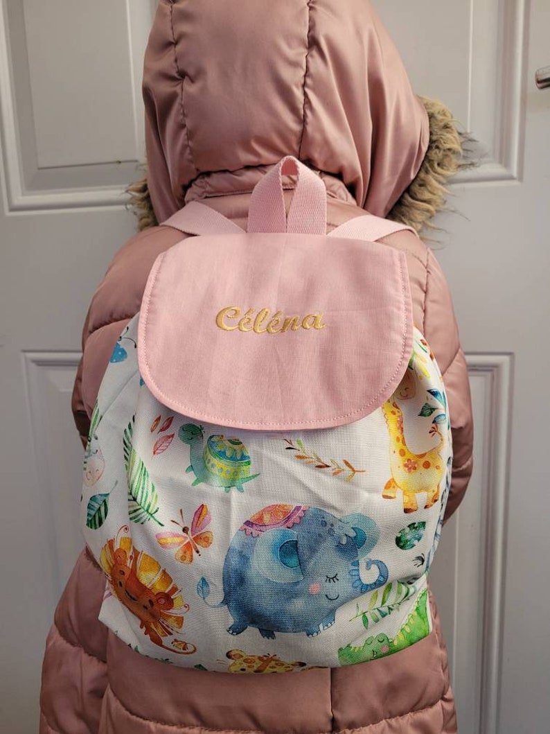 以下が含まれることがあります： ピンクと白のバックパックで、フラップには「Celena」という名前が刺繍されています。バックパックは白い背景にカラフルなジャングル動物のプリントが施されています。