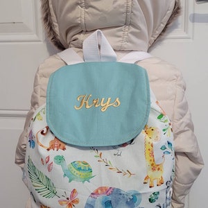 以下が含まれることがあります： 金色で「Krys」と刺繍されたフラップが付いた、薄い青と白のバックパック。バックパックは白い背景に、キリン、ゾウ、ライオン、カメなど、カラフルな動物のパターンが描かれています。