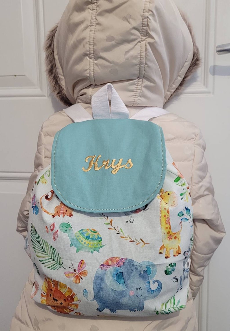 以下が含まれることがあります： 金色で「Krys」と刺繍されたフラップが付いた、薄い青と白のバックパック。バックパックは白い背景に、キリン、ゾウ、ライオン、カメなど、カラフルな動物のパターンが描かれています。