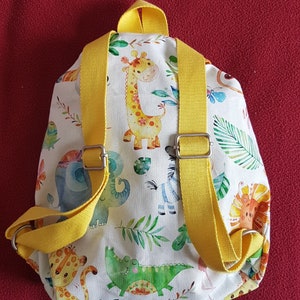 以下が含まれることがあります： 黄色いストラップが付いた小さな白いバックパック。バックパックは、キリン、ゾウ、シマウマ、ライオン、ワニなど、さまざまな動物のカラフルなプリントで飾られています。