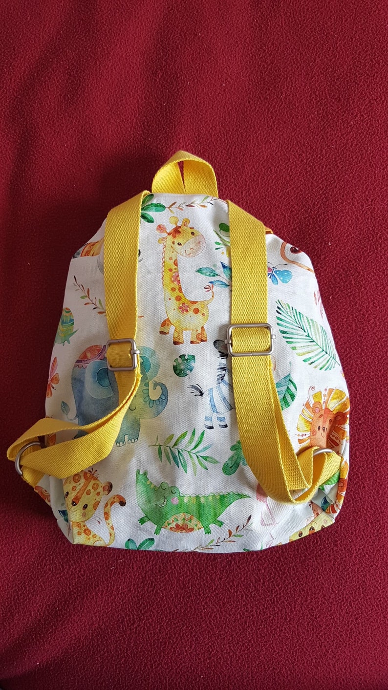 以下が含まれることがあります： 黄色いストラップが付いた小さな白いバックパック。バックパックは、キリン、ゾウ、シマウマ、ライオン、ワニなど、さまざまな動物のカラフルなプリントで飾られています。