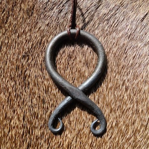 May include: A black metal pendant in the shape of a stylized knot, hanging from a brown leather cord. The pendant is on a brown furry surface.