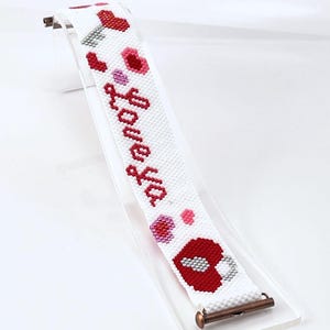 May include: White peyote stitch bracelet with the words "Love Ya" in red. The bracelet features red hearts and a kiss design. The bracelet is displayed on a clear stand with a copper clasp.