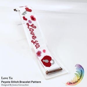 Könnte beinhalten: Ein weißes Peyote-Stich-Armband mit roten und rosafarbenen Herzmotiven und dem Schriftzug "Love ya" in roter Schreibschrift. Das Armband ist an einem Ende mit einem metallischen, kupferfarbenen Stangenverschluss versehen und zeigt ein Herzmuster.