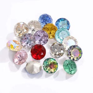 Pode incluir: Conjunto de strass redondos e facetados de vidro em várias cores, incluindo transparente, azul, verde, vermelho, roxo e dourado. Os strass estão dispostos sobre uma superfície branca.