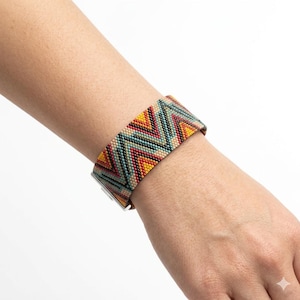 Pode incluir: Uma pulseira larga de contas com um padrão de chevron em tons de amarelo, vermelho, verde-azulado e bege. A pulseira é presa com um fecho prateado. O design é um padrão geométrico colorido.