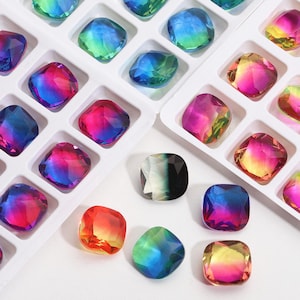 May include: A collection of square, faceted glass cabochons featuring a vibrant gradient of colors including blue, purple, pink, yellow, and green. Some stones are arranged in a white plastic grid tray, while others are scattered loosely.