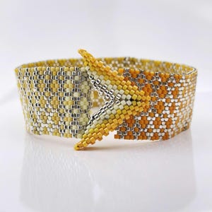 Puede incluir: Una pulsera ancha de cuentas tejidas con un patrón geométrico en tonos amarillo, naranja y plata metálica. La pulsera presenta un diseño de cierre superpuesto en forma de punta o chevron, creado con pequeñas cuentas cuadradas uniformes.
