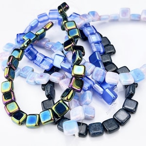 May include: A collection of square-shaped glass beads strung together, featuring a variety of colors including iridescent metallic blue, deep navy, and translucent pale lavender, arranged in a tangled cluster against a plain white background.