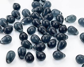 May include: A collection of small, teardrop-shaped beads in a translucent dark gray color. Each bead features a smooth, polished surface with a visible hole for threading, scattered across a plain white surface in a loose, unorganized arrangement.