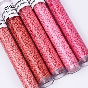 May include: Five clear tubes filled with small, pink, square-shaped beads. The beads vary in shades of pink, from light to dark. Each tube has a white label with black text. The beads are opaque and appear to be made of glass or plastic.