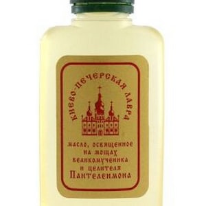 May include: A small, clear glass bottle with a green lid. The bottle contains a light yellow liquid. The label on the bottle is gold with red text and depicts a church. The text on the label reads "Киево-Печерская Лавра" and "масло, освященное на мощах великомученика и целителя Пантелеимона".